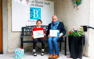 schoolgirl and resident sat on bench holding winning drawing and certificate