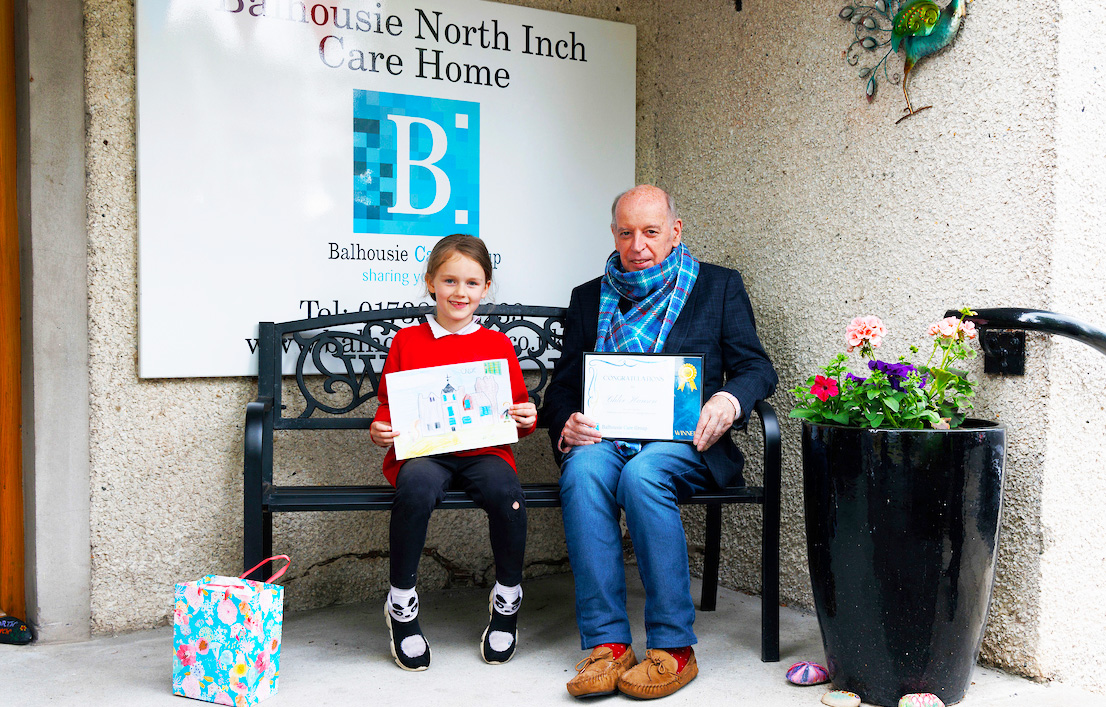 schoolgirl and resident sat on bench holding winning drawing and certificate